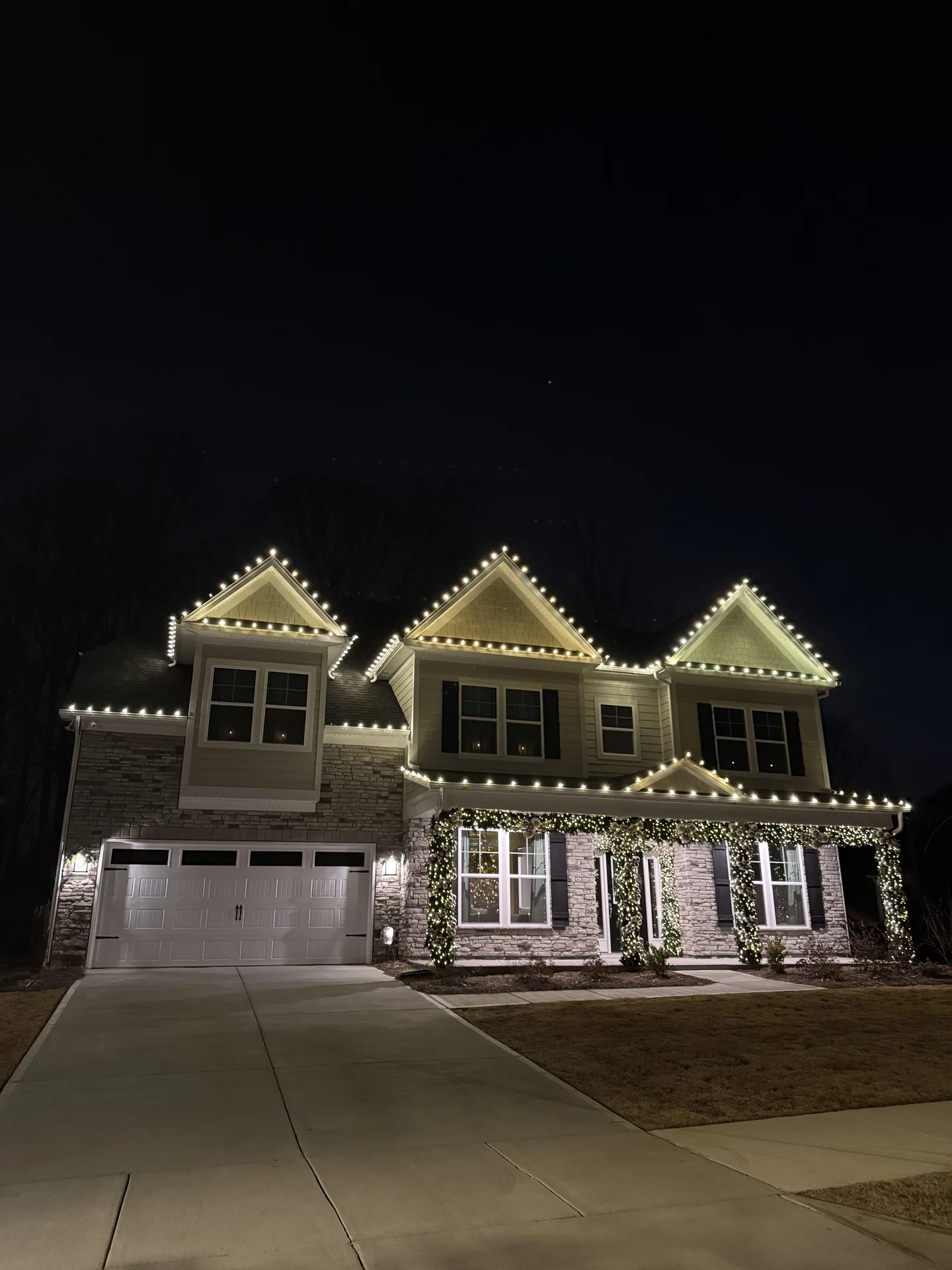 Multi-story home outlined with warm white roofline Christmas lights and illuminated evergreen entry decorations at night