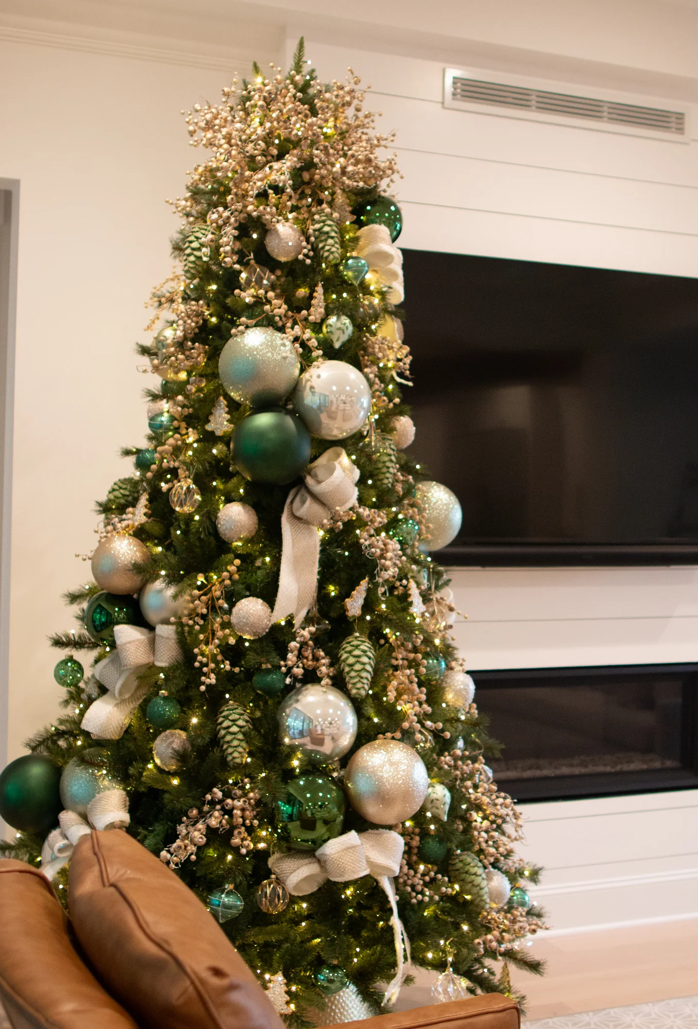 Full Christmas tree decorated with emerald, ivory, and champagne ornaments and warm lights beside a white fireplace