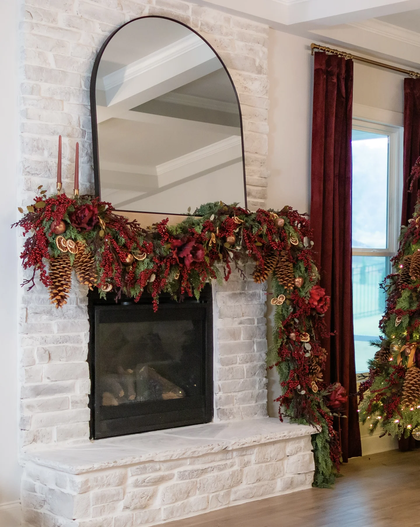 White brick fireplace mantel decorated with burgundy florals, berries, pinecones, and cascading garland